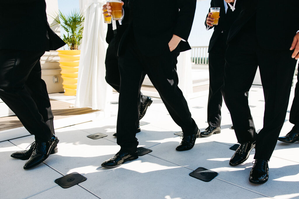 Groomsmen walk across black and white tile floor along the pool with beers in their hands