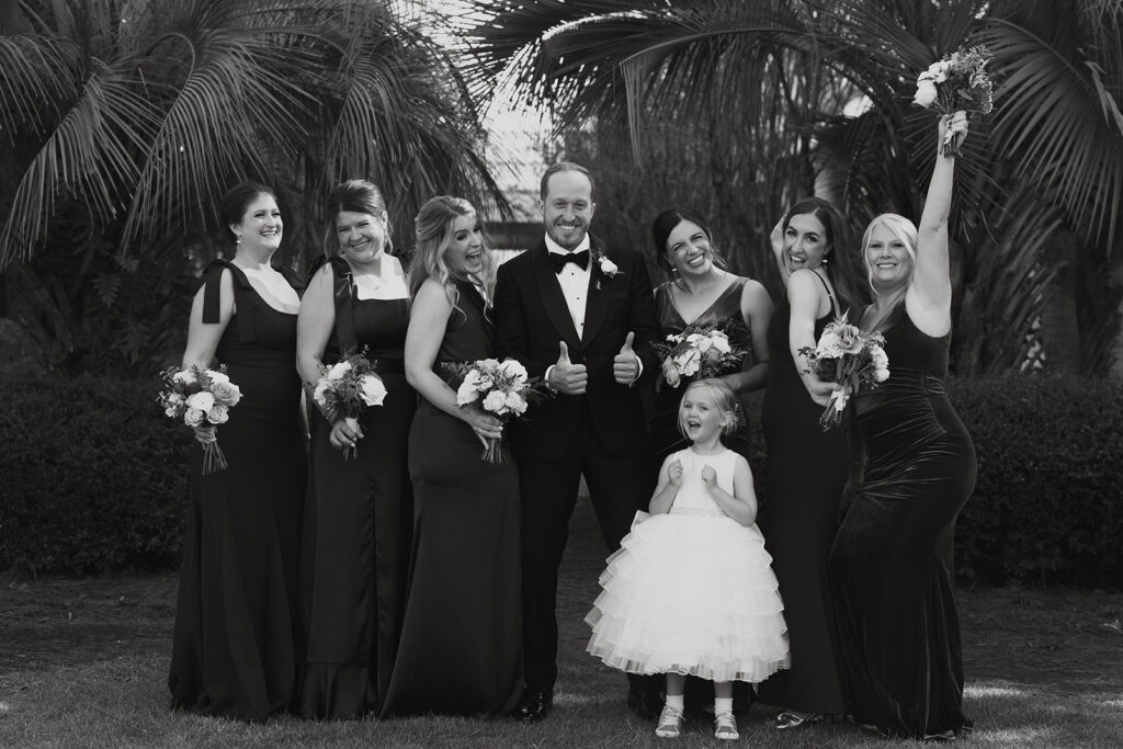 Groom poses with two thumbs up with bridesmaids and flower girl surrounding him