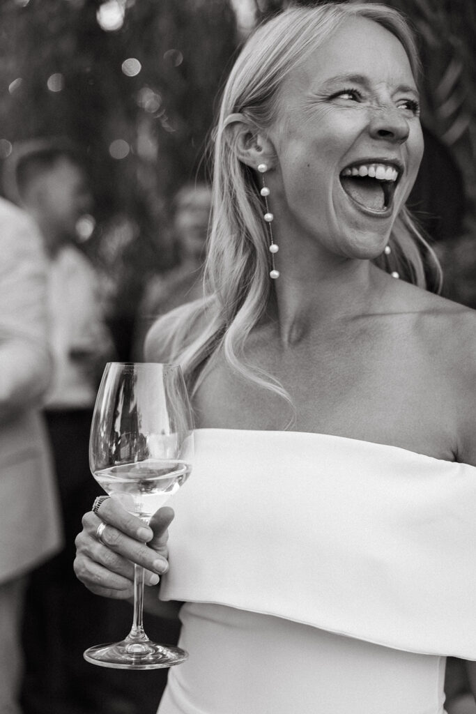 Bride looks of camera with ebullient smile while holding a non-alcoholic glass of wine.