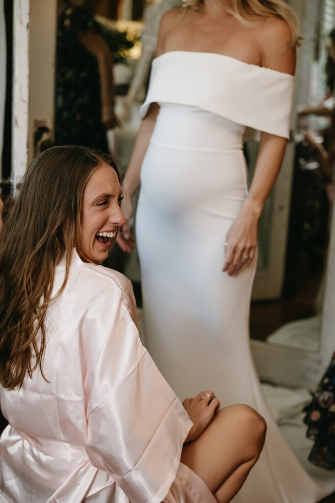 Maid of honor grins as she helps bride put her shoes on