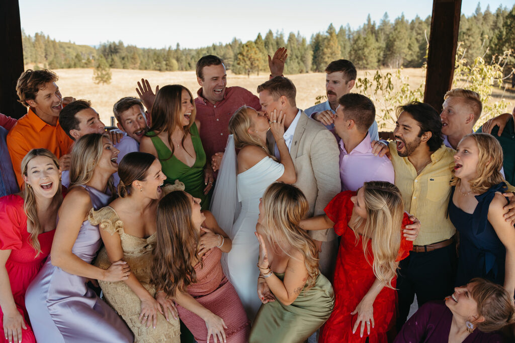 Colorful bridal party looks on as bride and groom kiss.