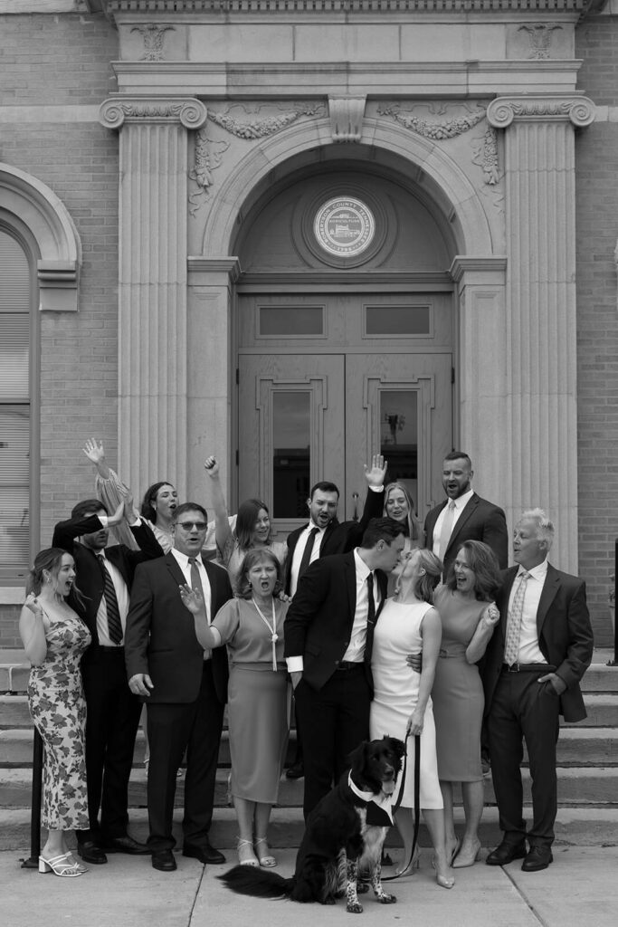 Bride and groom kiss while their intimate guest list celebrates behind them on courthouse steps.