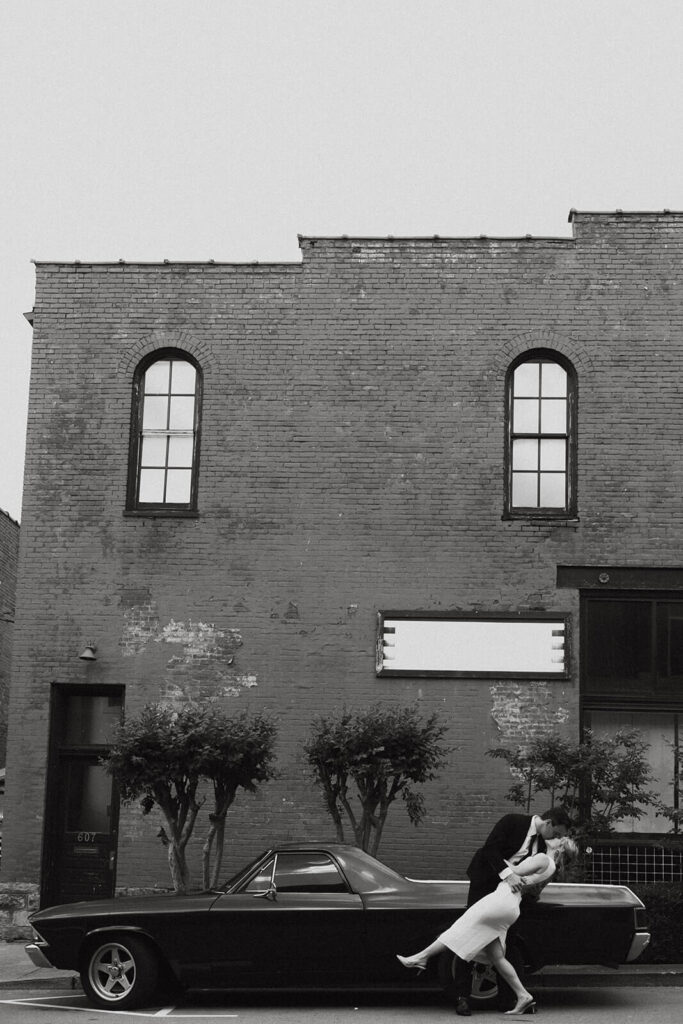 Groom dips bride in front of vintage car in classic photograph