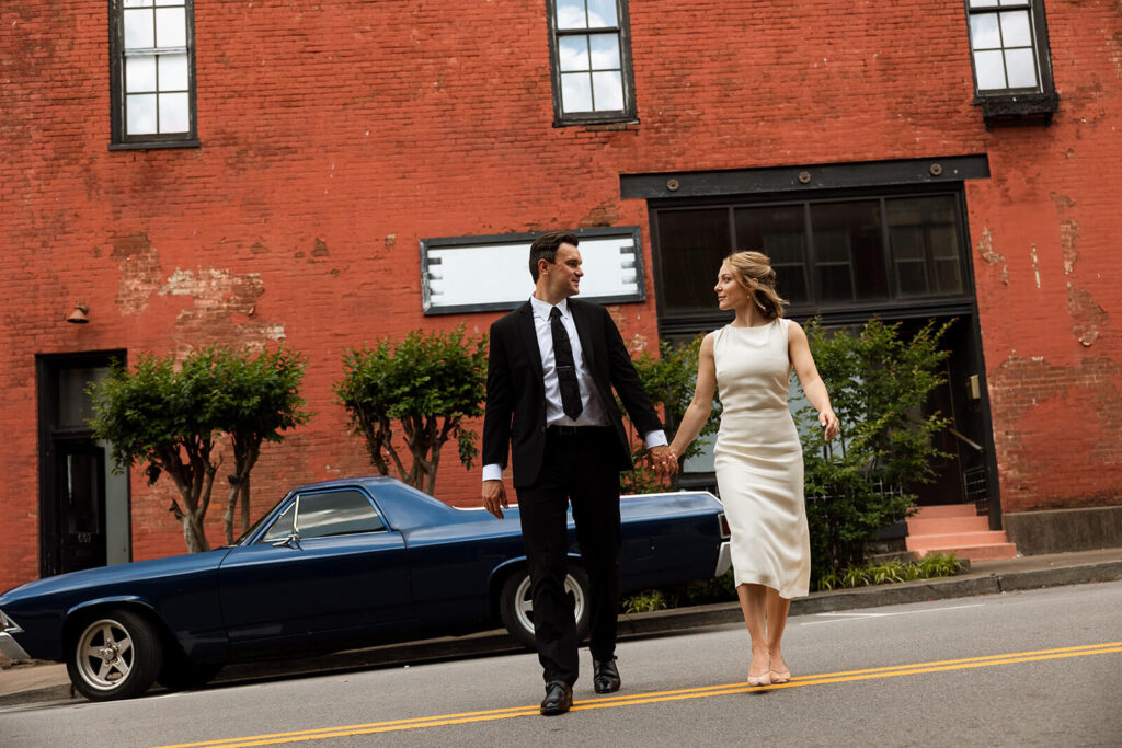 Bride and groom cross Main Street hand in hand with vintage navy car in the background