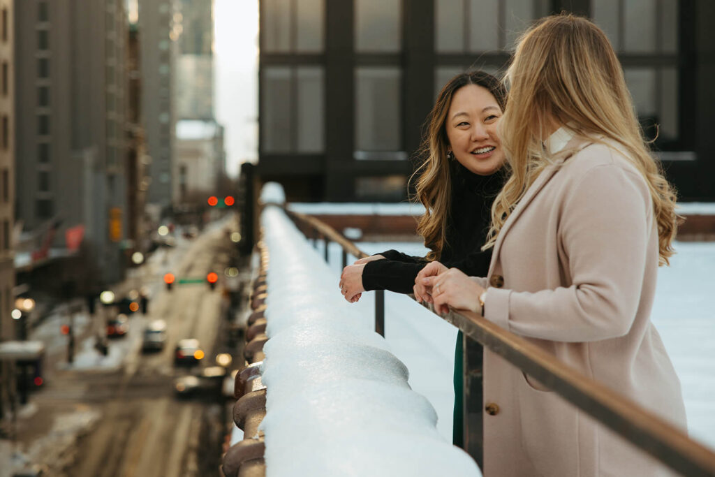 Engaged couple smiles at the edge of a Nashville rooftop overlooking the snowy street below