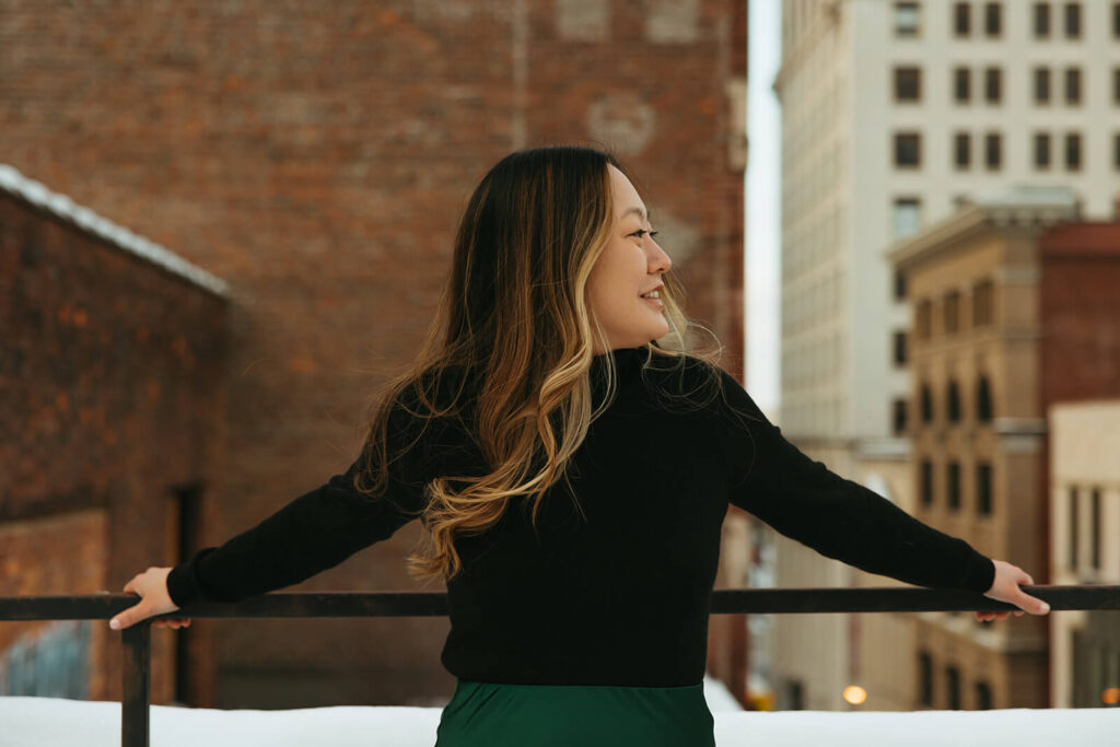 Woman in black turtleneck sweater and emerald green skirt leans against rooftop balcony in Nashville engagement photoshoot. 