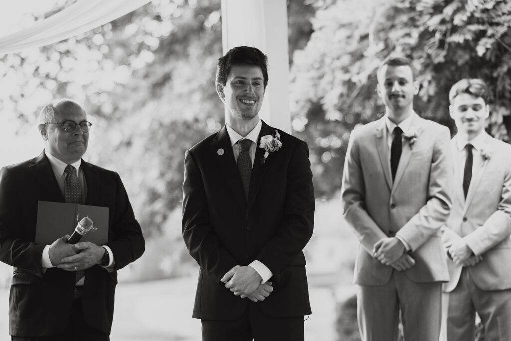 Groom grins at his bride as she comes down the aisle towards him in documentary style wedding photos