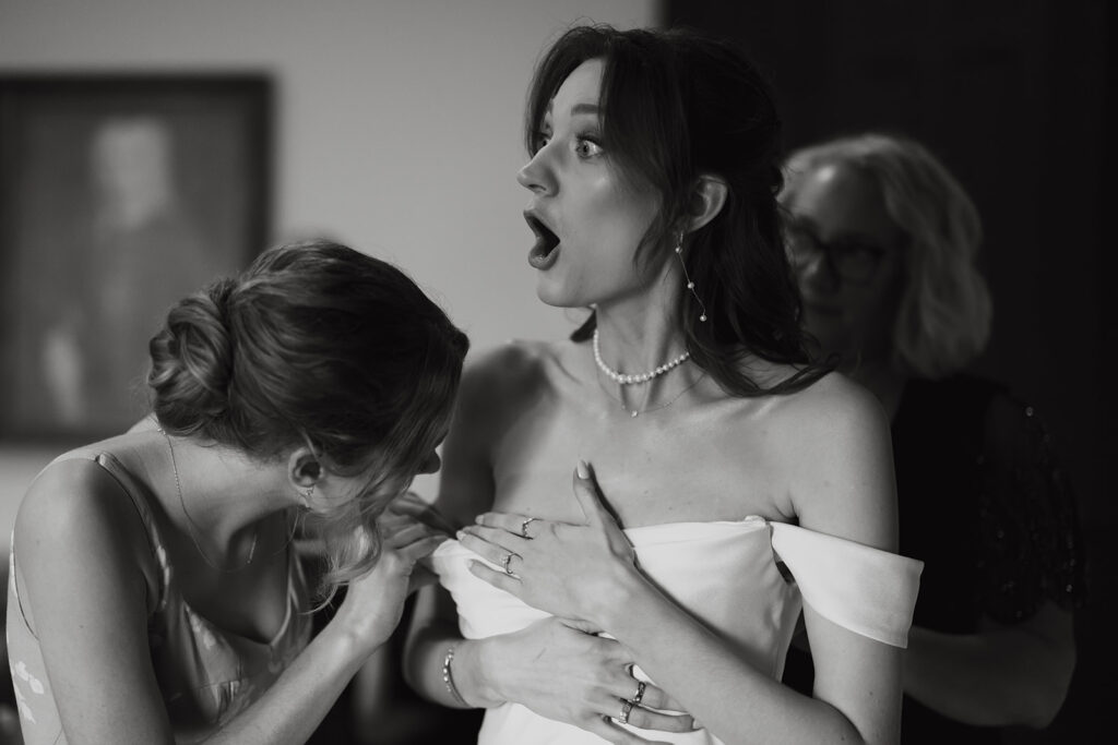 Bride reacts with surprise to something a bridesmaid said as her dress is being buttoned up