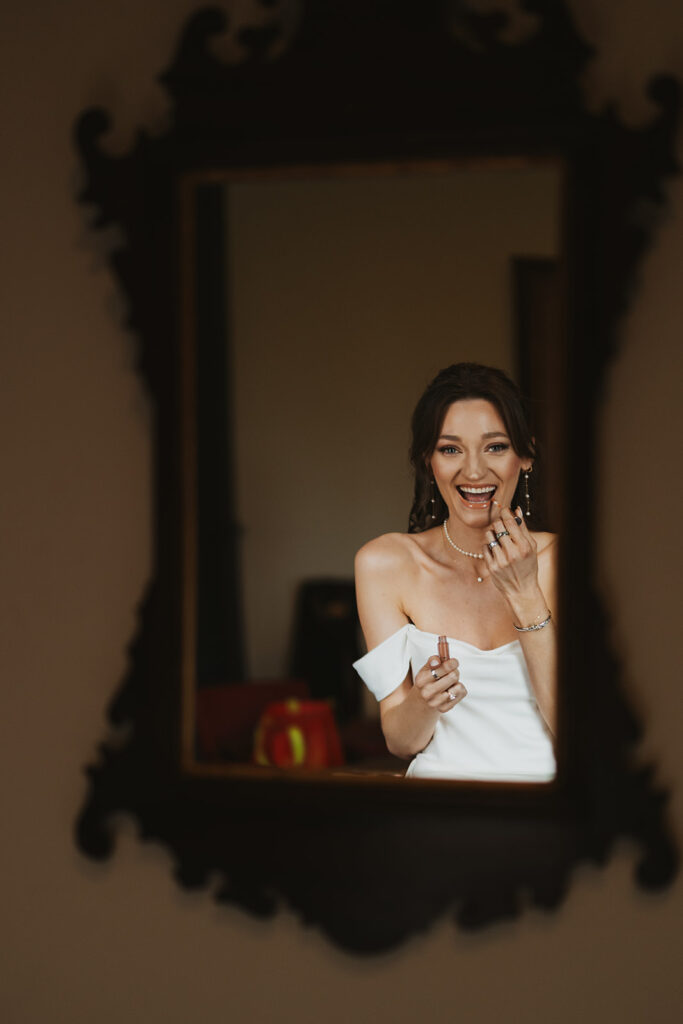 Bride grins at documentary style wedding photographer in the mirror while applying her lipstick