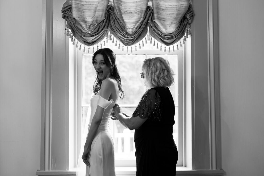 Bride gives the camera an open-mouthed grin of excitement as mother of the bride buttons up her dress