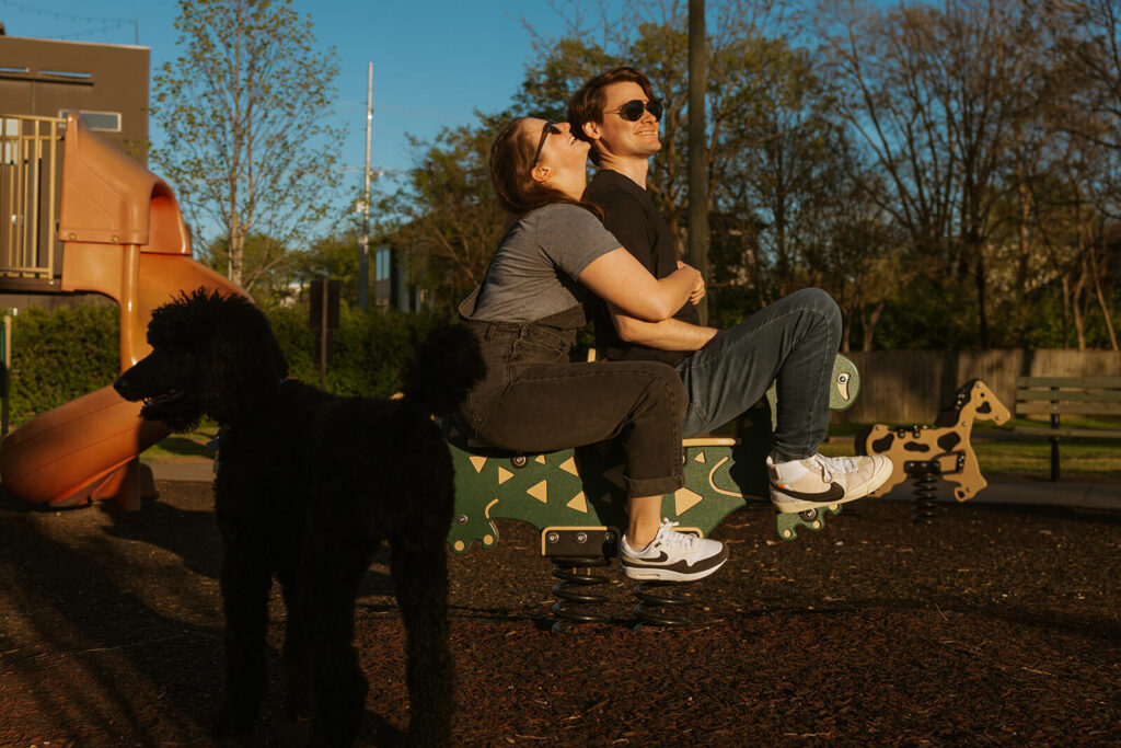 Couple plays on the playground while black poodle hangs out nearby