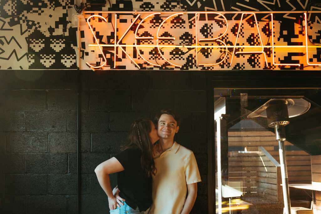 Engaged couple poses under neon Skeeball sign