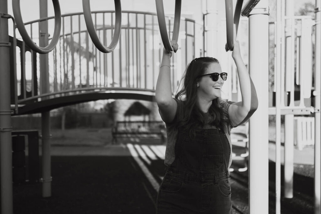 Women wearing sunglasses and overalls smiles at her fiancee while holding on to monkey bars