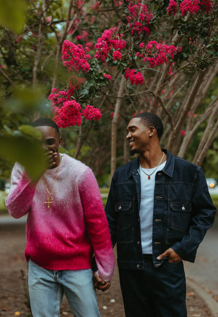 Man in pink sweater covers his face in laughter while his fiance looks on adoringly