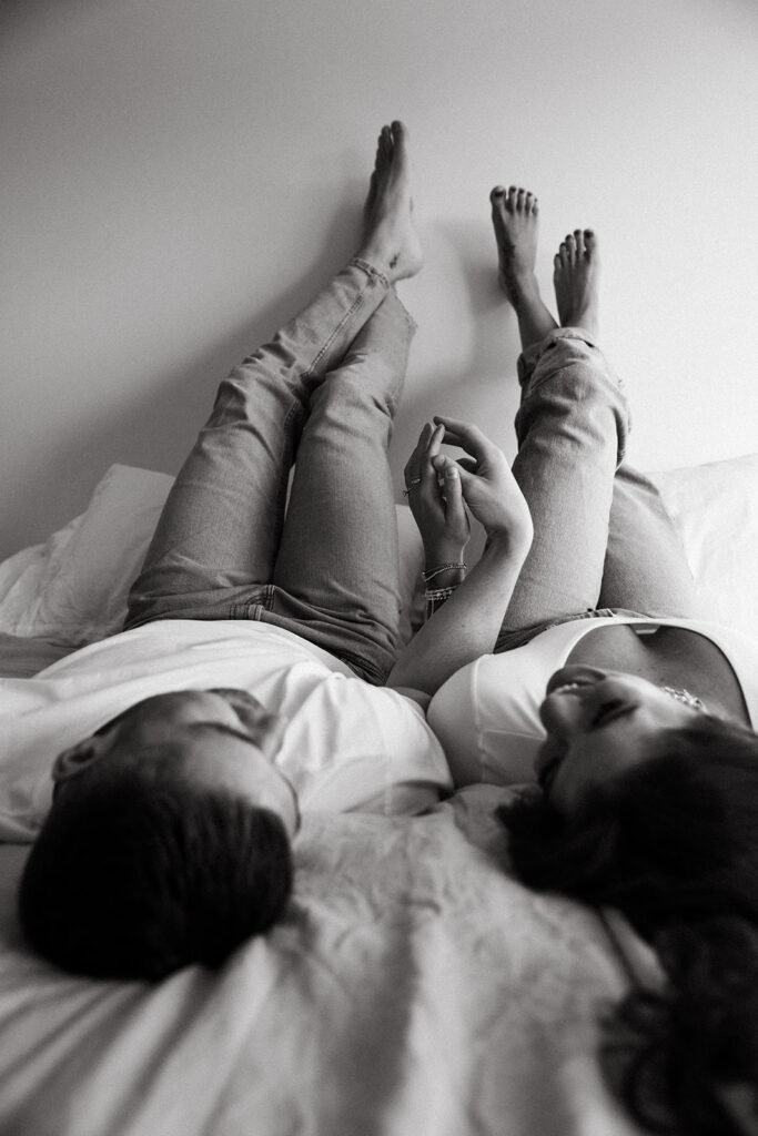 Couple intertwines hands as they lay in bed with their feet up the wall