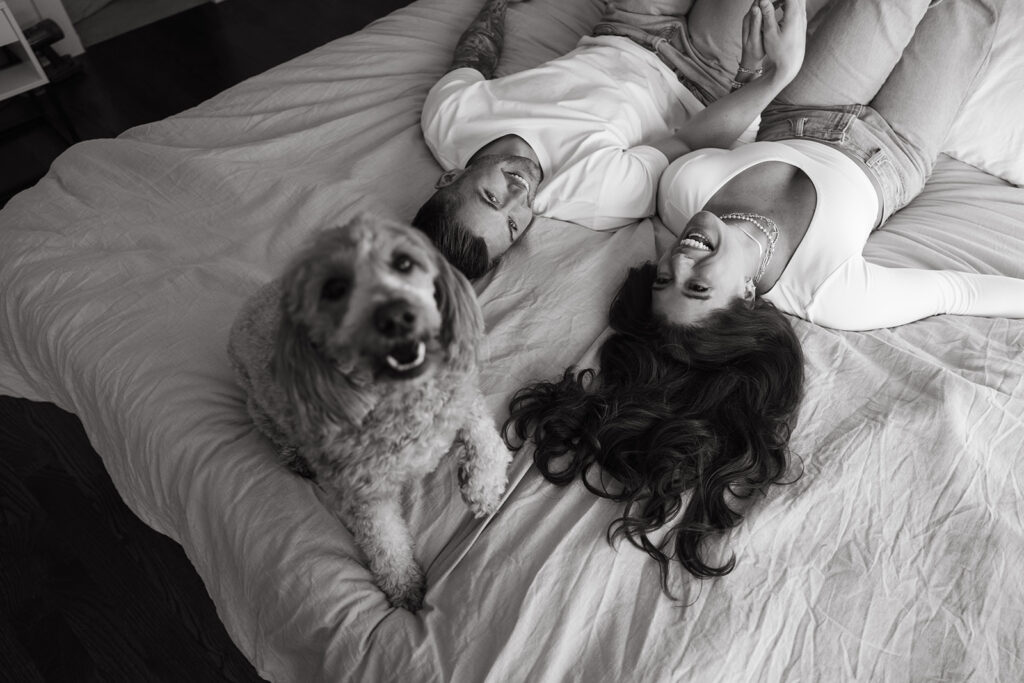 Engaged couple lays on bed while their dog looks up at the ceiling