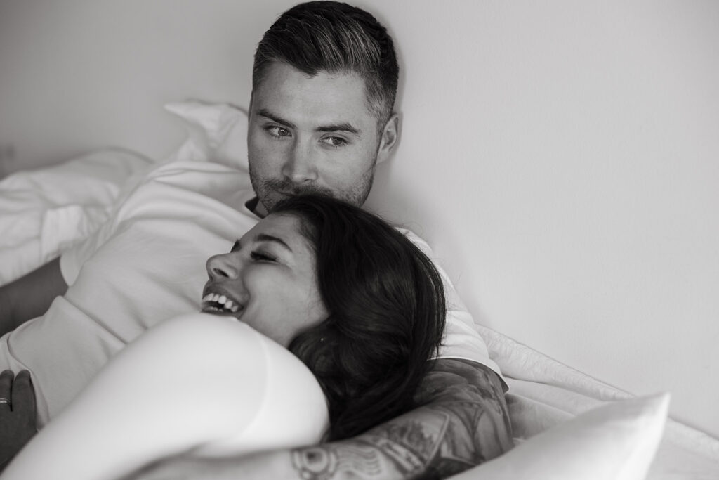 Woman laughs as she lays her head on her fiance's shoulder while they're laying in bed