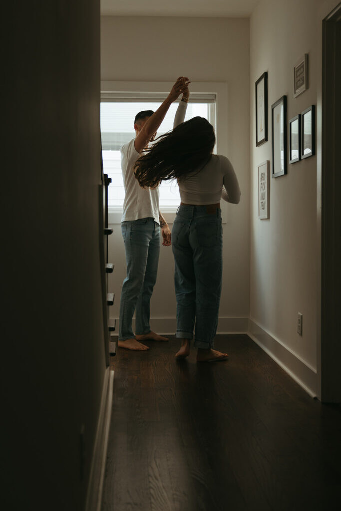 Man in denim jeans and white tee twirls his fiance around in their hallway