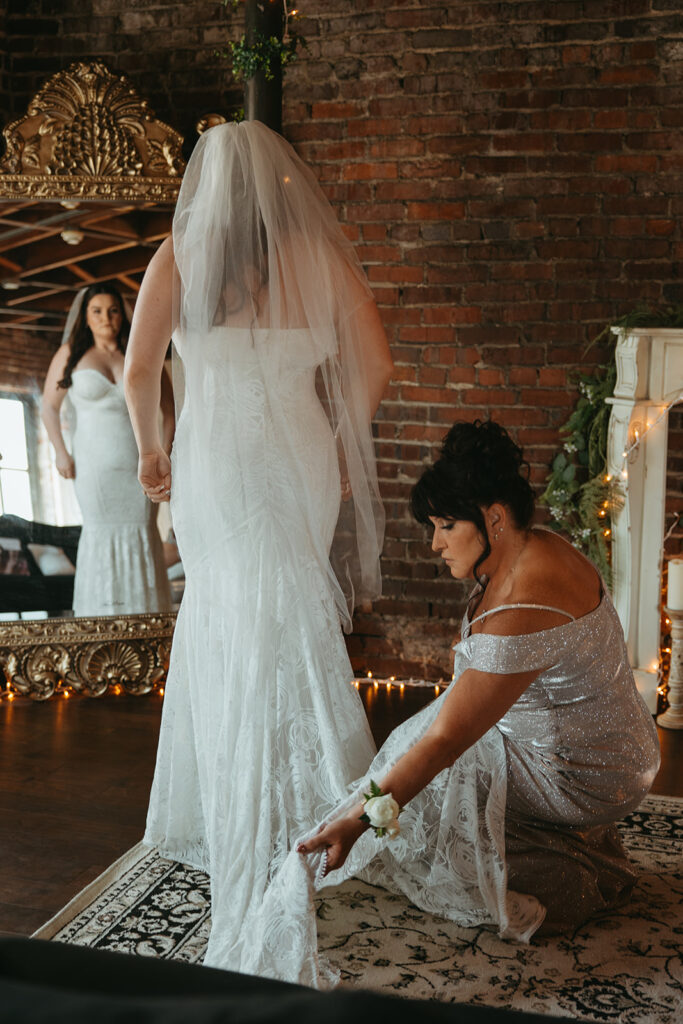 Mother of the bride fixes train while bride admires her reflection in the vintage gold mirror