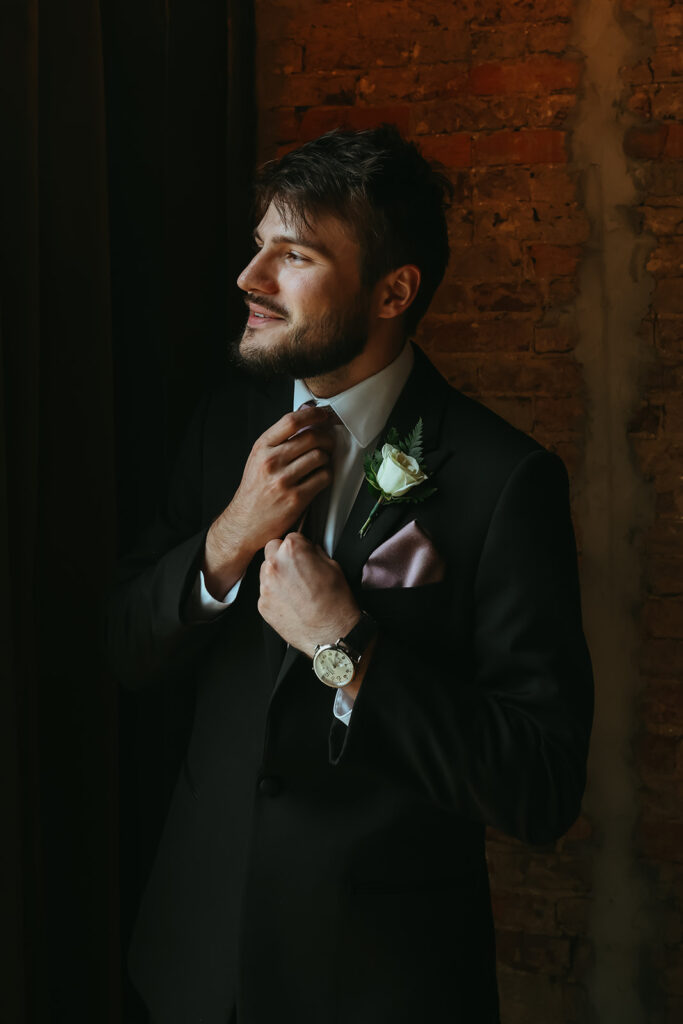 Groom looks out the window as he fixes his tie