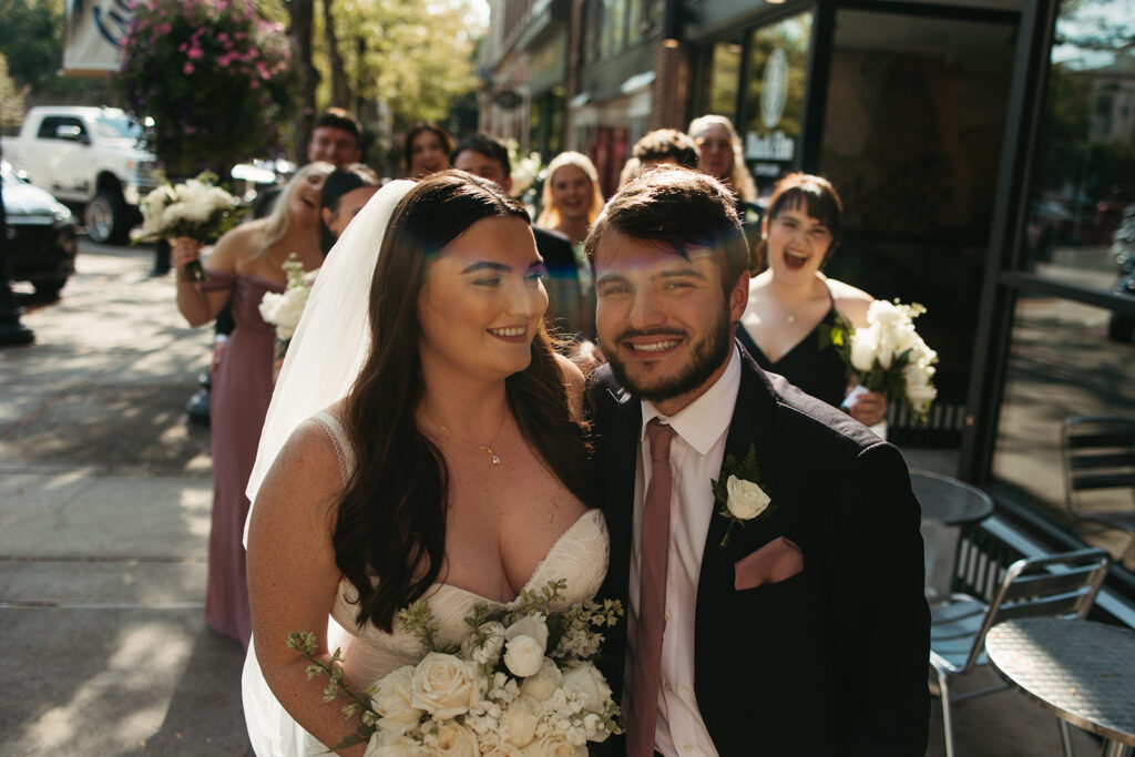 Bride smiles at groom under direct sunlight while wedding party traipses behind them