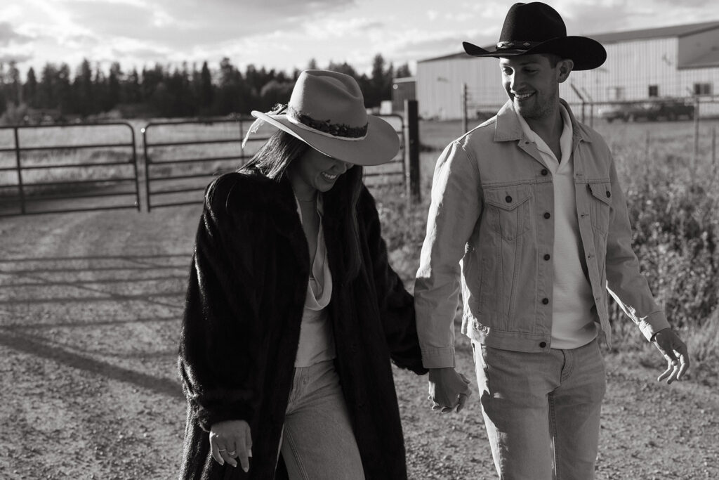 Woman wearing fur coat and man in denim walk down gravel trail hand in hand.