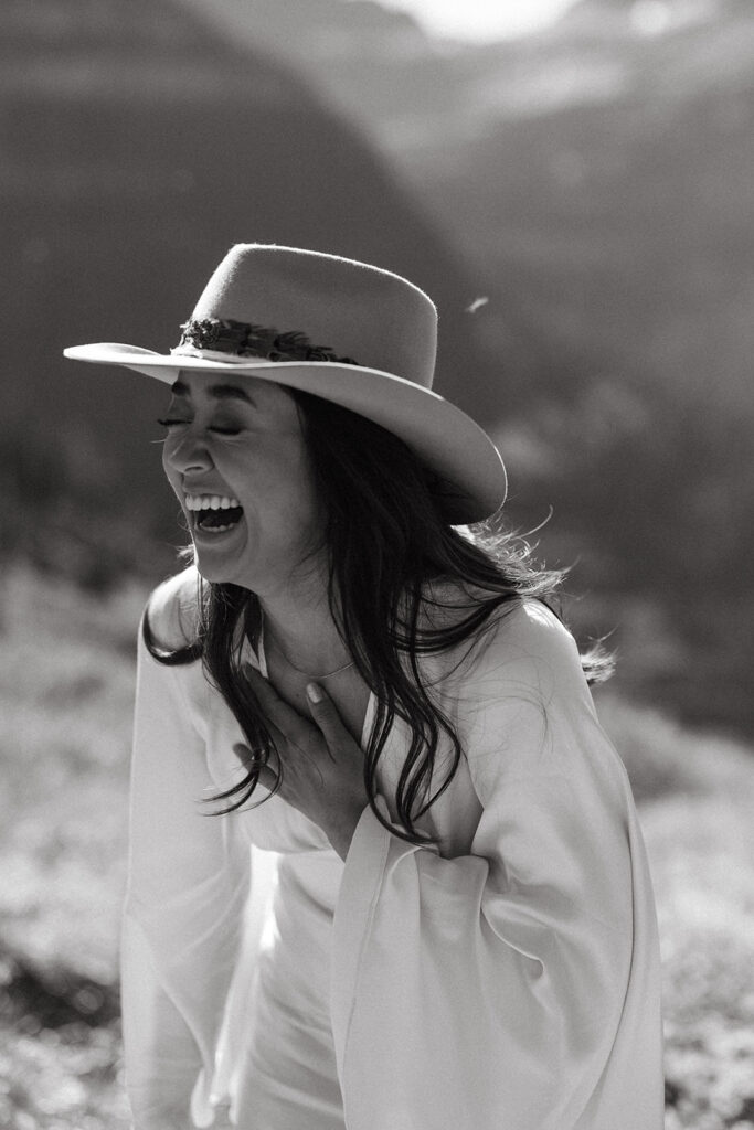 Bride wearing cowboy hat and wedding gown laughs as she stands in the mountains of Glacier National Park.