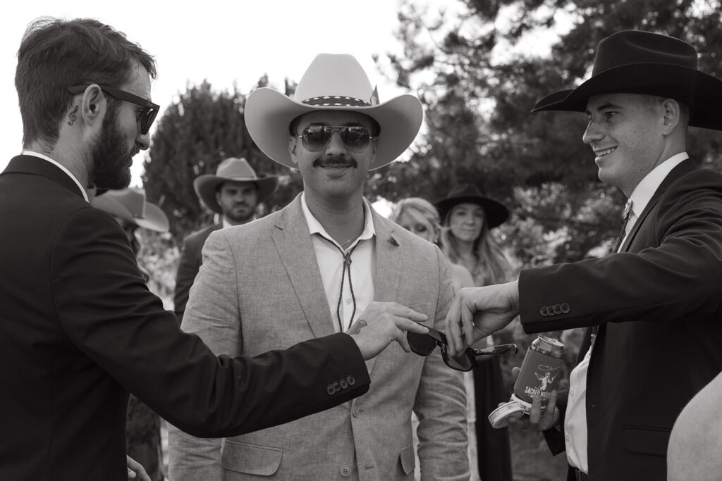 Wedding guests pass off sunglasses to each other as another wedding guest smiles at the camera with his mustache and cowboy hat. 