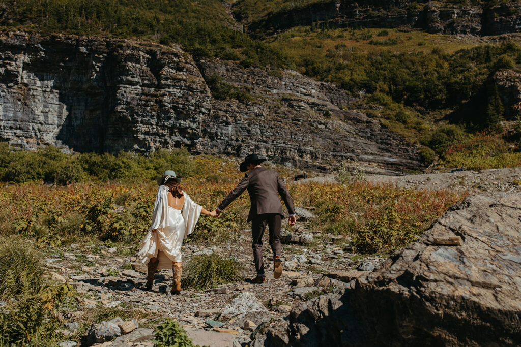 Groom holds bride's hand as she walks over rocks in Glacier National Park. 