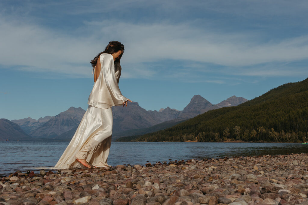 Bride walks barefoot along shoreline of glacial waters in Glacier National Park in Montana. 