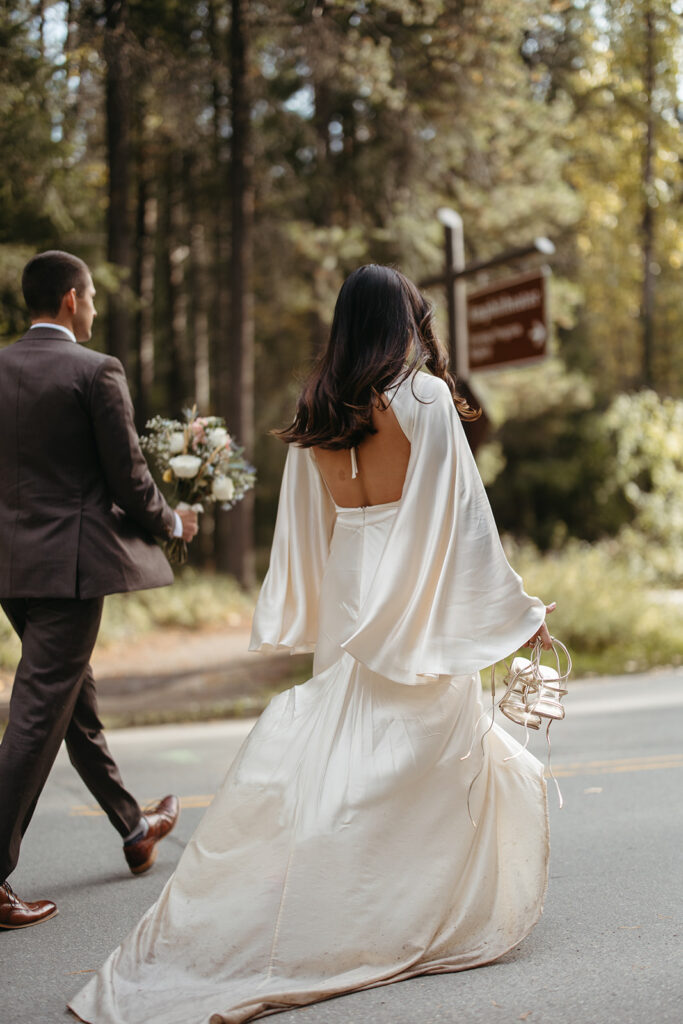 Bride carries her shoes and groom carries bridal bouquet as they walk toward the car after their Yellowstone wedding ceremony. 