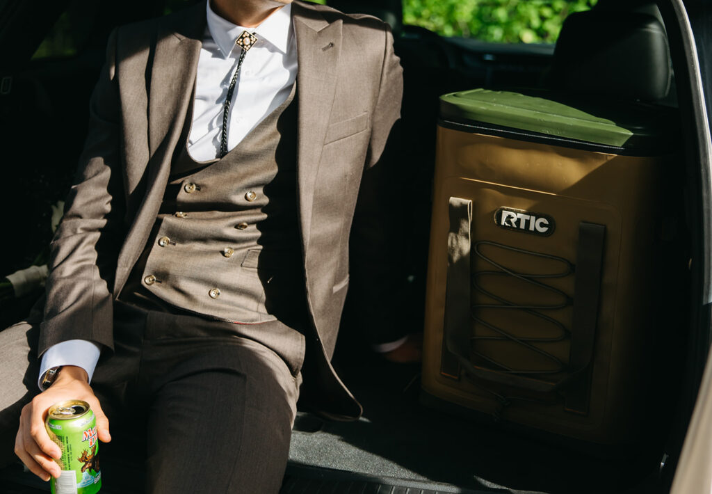 Groom sits in trunk of car next to RTIC cooler after his wedding ceremony. 