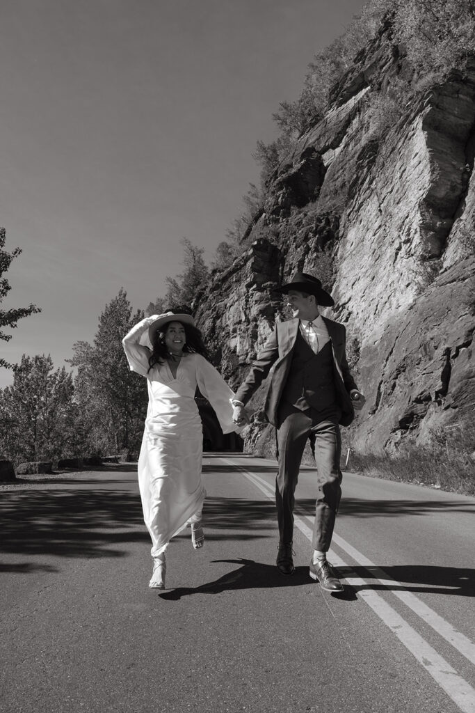 Bride holds onto her cowboy hat as she runs hand in hand with her groom on Montana highway. 