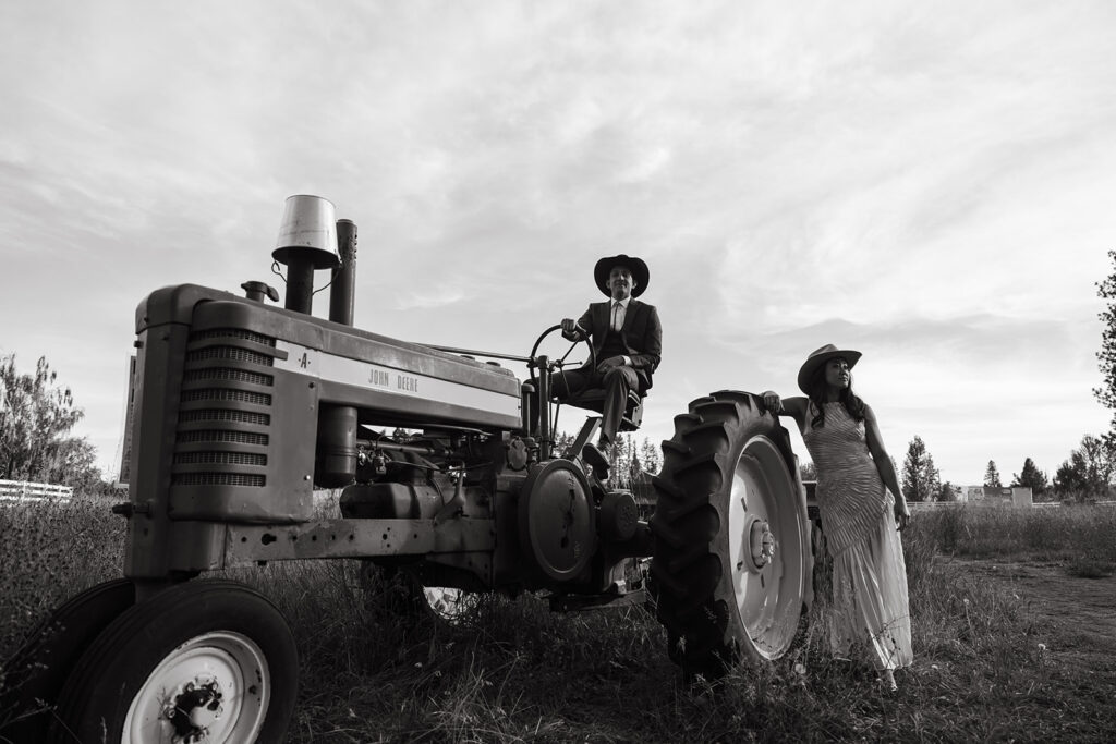 Groom sits on tractor while bride props her arm on the wheel. 