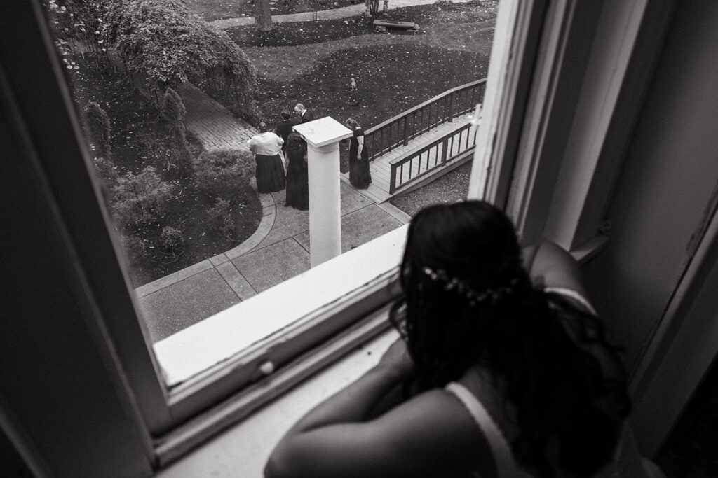 Bride peeks out of upstairs window as wedding guests start to arrive