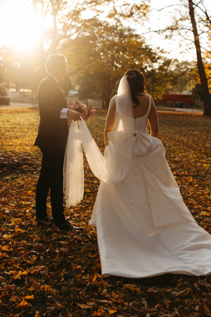 Groom holds bride's veil off the ground to keep it from gathering leaves during autumnal black tie wedding in Nashville