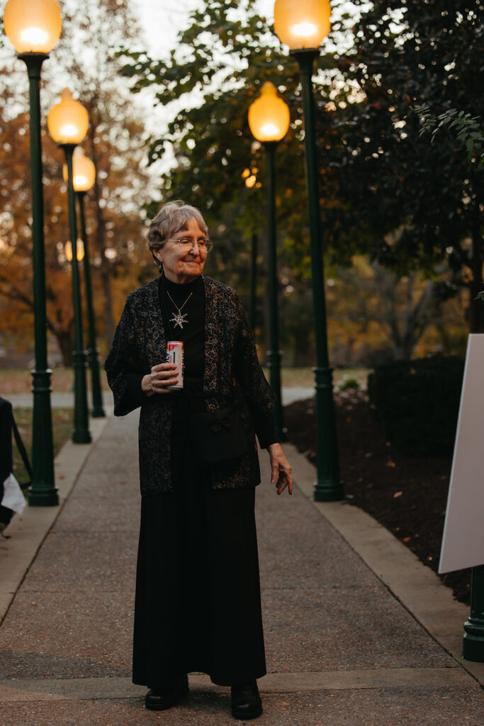 Elderly woman shows of her black tie wedding attire while lamplights turn on behind her