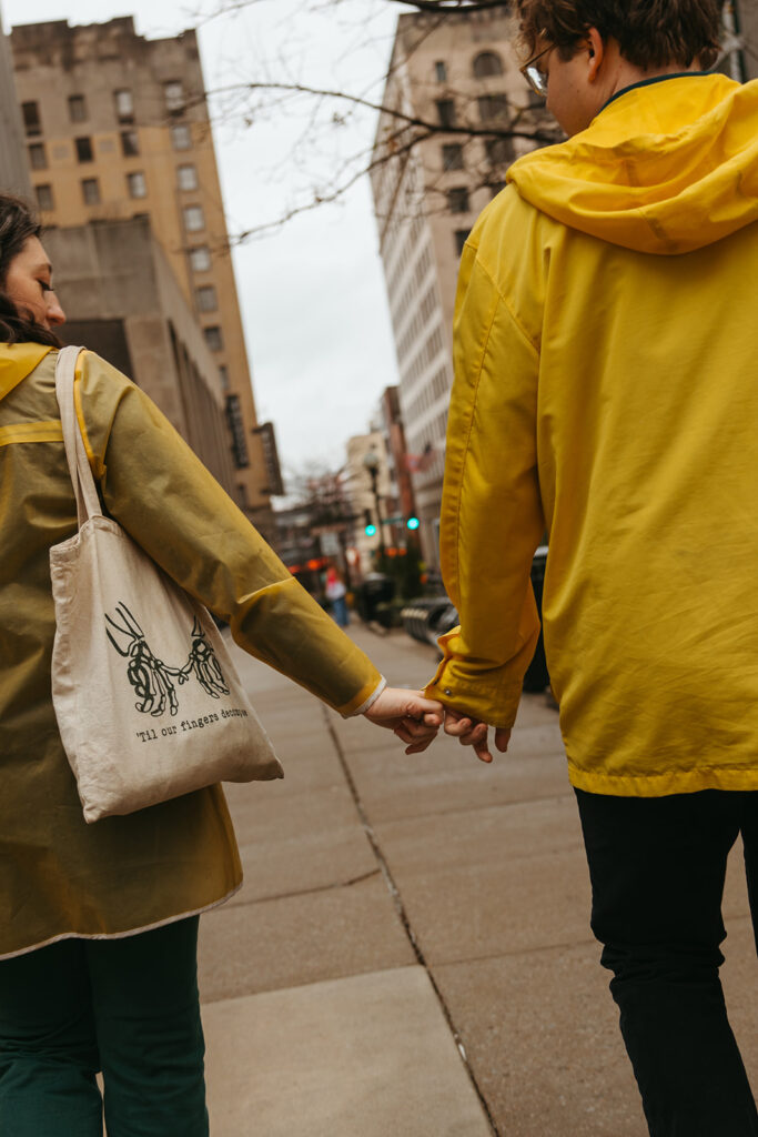 Engaged couple walks through the streets of rainy Nashville with their fingers intertwined, mirroring the Noah Kahan 'Everywhere, Everything' tote that the woman is carrying.