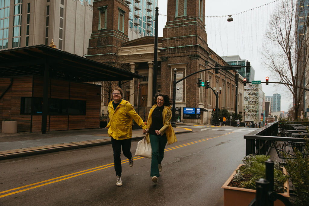 Playful and quirky engaged couple runs through the streets of downtown Nashville during a downpour.