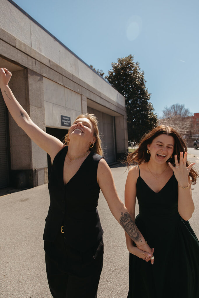 Woman in black pantsuit gleefully raises her fist in the air while skipping hand in hand with her fiance