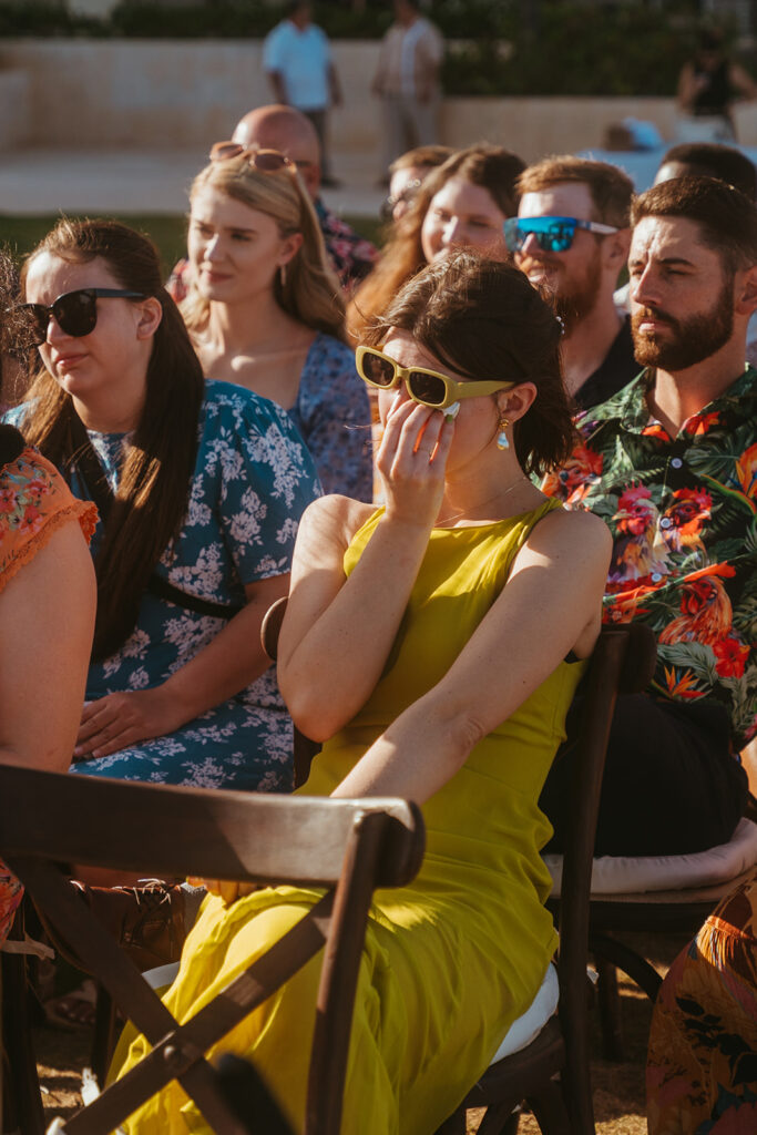 Wedding guest wearing a lime green dress and sunglasses wipes a tear at destination wedding in Cancun
