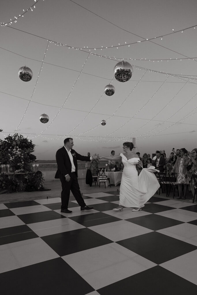 Bride and groom share first dance on black and white checkered floor with disco balls and string lights overhead
