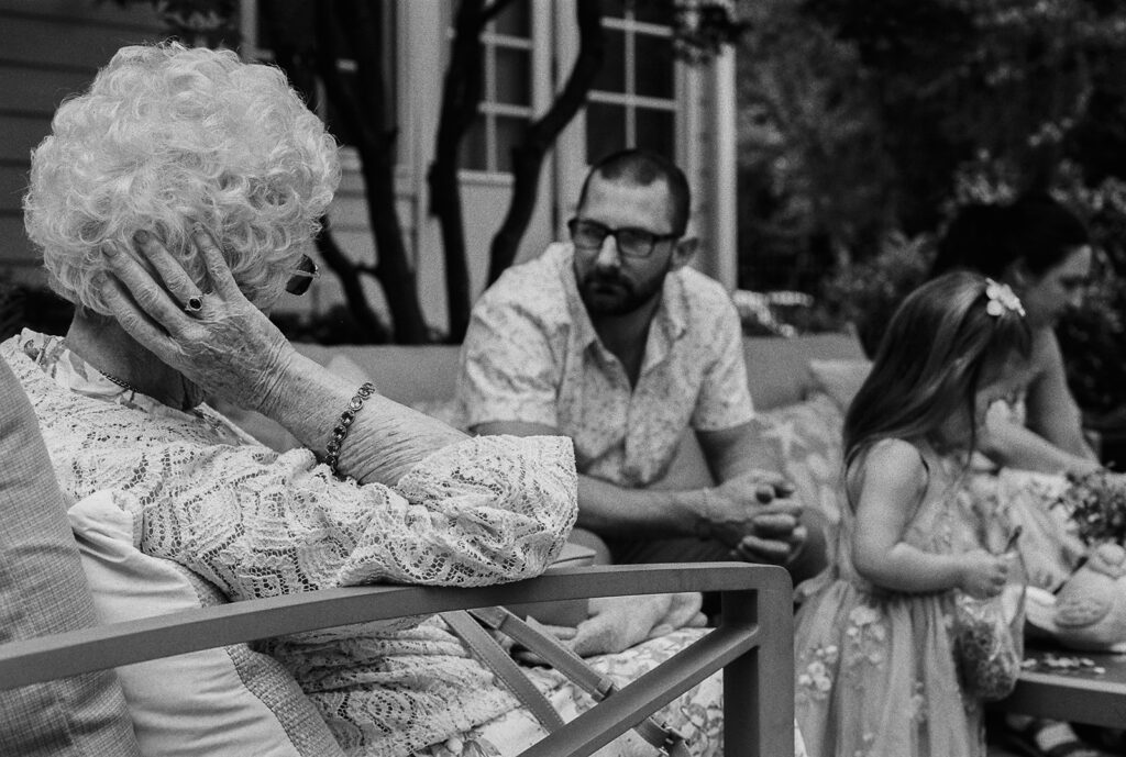 Grandmother sitting in a chair during the backyard wedding reception while guests chat nearby