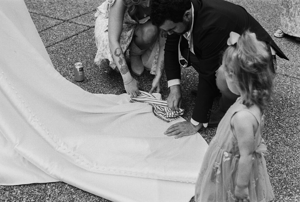 Guests kneeling to blot the hem of the bride’s dress as a flower girl watches at this laid-back outdoor wedding