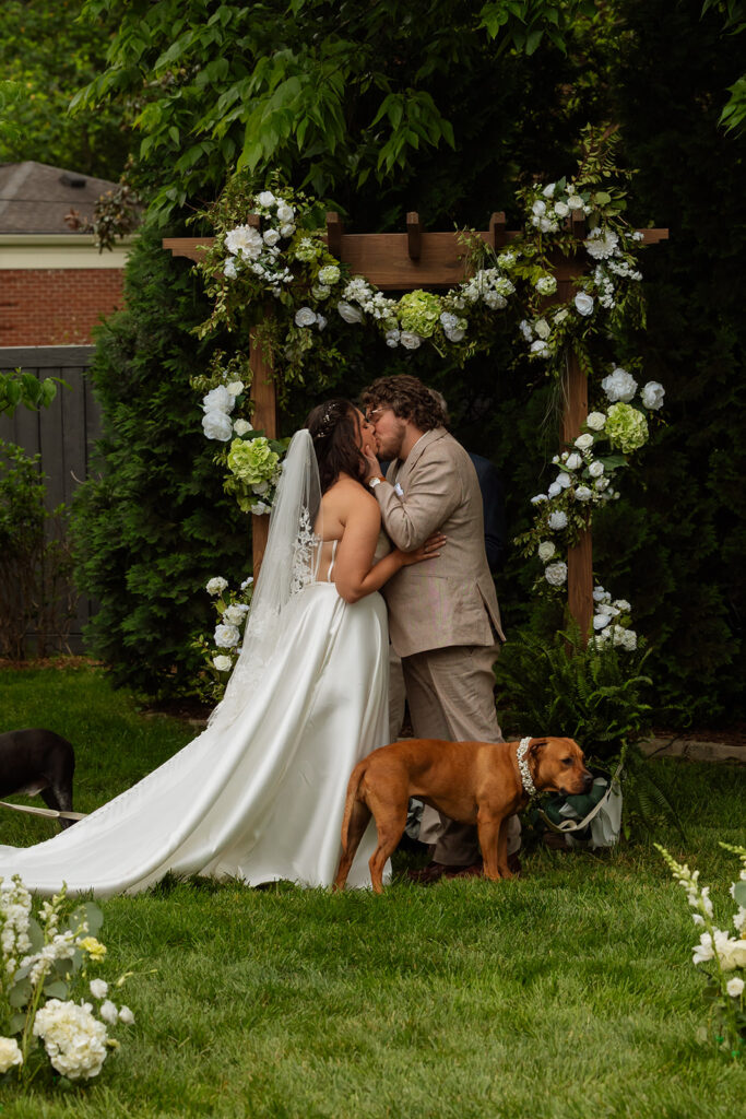 Couple sharing a kiss under the floral arbor with their dog at their backyard wedding ceremony
