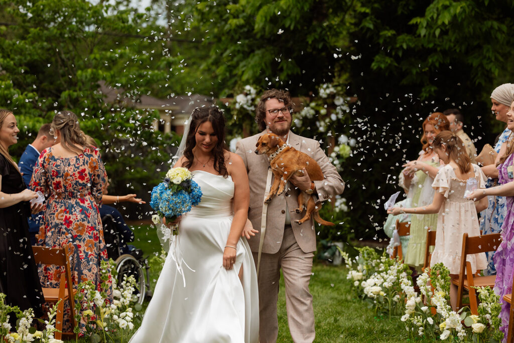 Guests cheering as the couple exits the backyard wedding ceremony with their dog in hand

