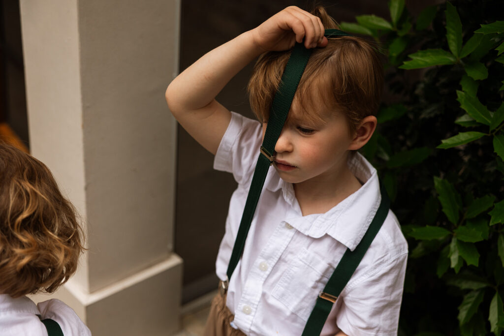 Young boy in suspenders adjusting his strap before the ceremony at a playful backyard wedding