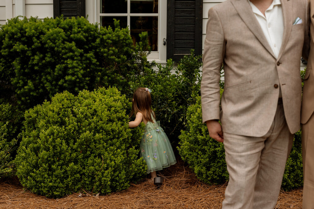 Flower girl in a green dress exploring the bushes before the backyard wedding ceremony