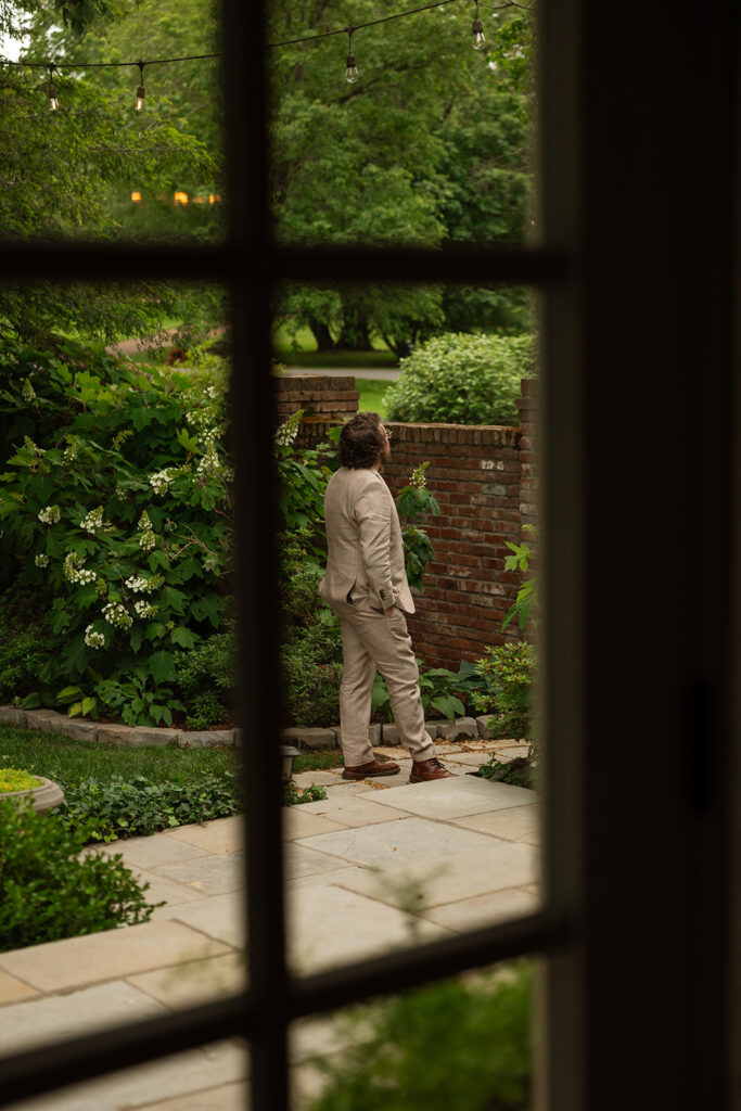 Groom standing in the garden while he waits for bride, viewed through a window of at-home wedding