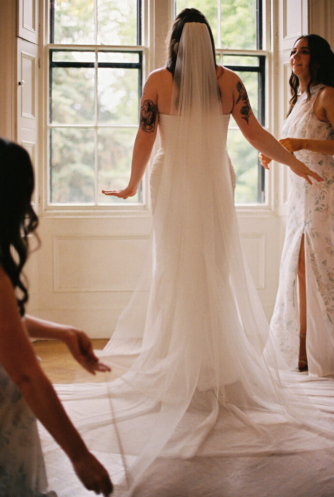 Bride standing at a tall window in her veil as bridesmaids help arrange the train of her dress.